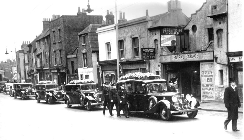 View of funeral cars, three with floral tributes, accompanied by 6 men from the Fire Station in uniform