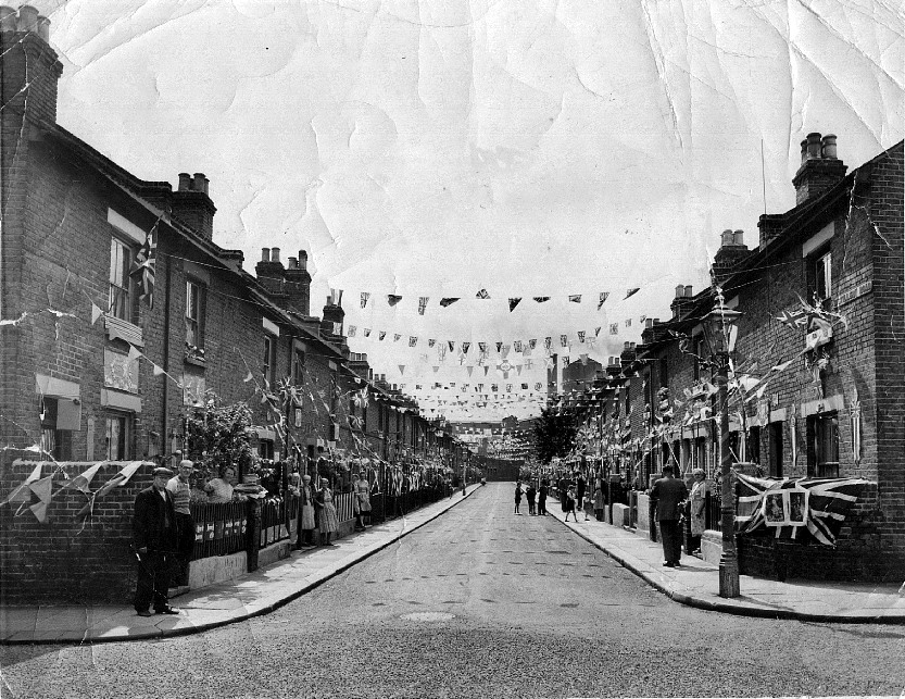View along street festooned with bunting and union jacks, several residents standing in their front gardens