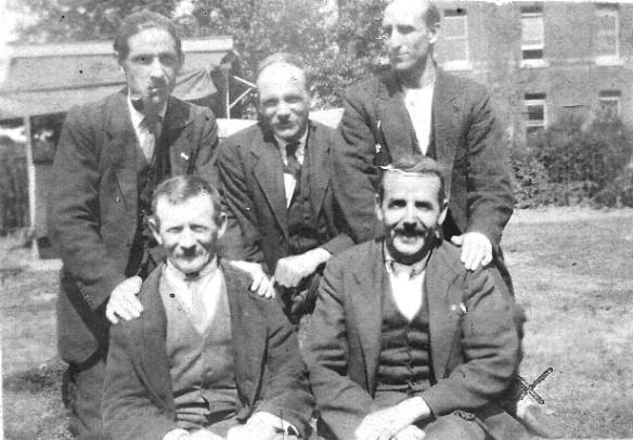 Five men posed for photo in a garden, large brick house in background; they are wearing waist coats and jackets and by their relaxed appearance, closeness, hands on shoulders, appear to know each other well